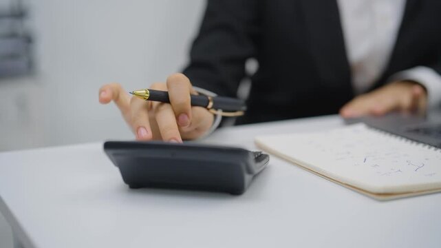 Businessperson in formal suit using calculator and taking notes with pen on notepad at white desk in office environment