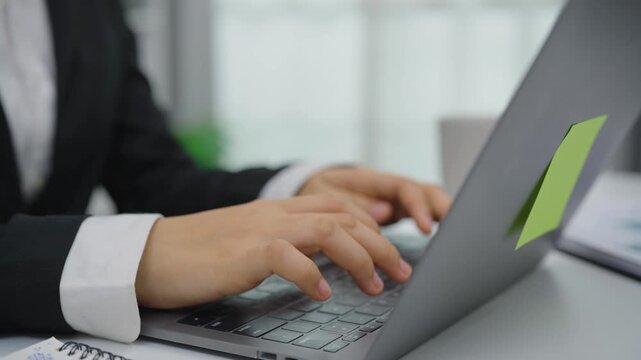 Business person wearing suit typing on laptop keyboard working on project in modern office environment with notebook