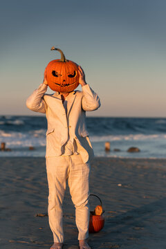 Person wearing a carved pumpkin on a beach at sunset. A person in a suit holds a carved pumpkin over their head on a beach at sunset.