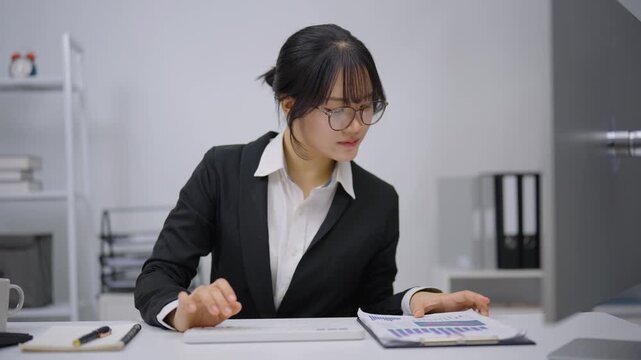 Young professional woman in glasses and business suit sitting at desk with computer analyzing documents in modern office