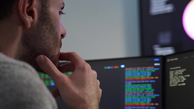 Software developer thinking with hand on chin while reviewing programming code on multiple computer monitors, reviewing coding script running data