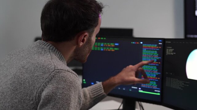 Software engineer points at lines of script programming code on a computer monitor while analyzing software at a multi screen at the office