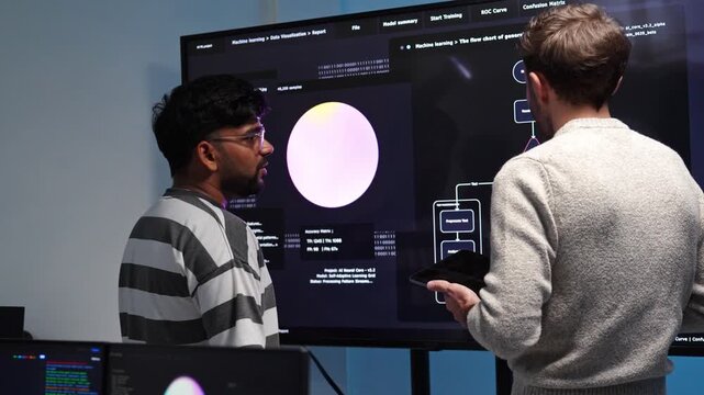 Software developer discuss machine learning data on a large screen, one holding a tablet while reviewing AI assistant model training results and a flow chart in a tech office.
