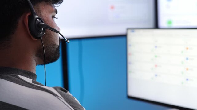 Indian male technical support wearing a headset looks at multiple computer monitors displaying data dashboards in tech office, solving customer problem
