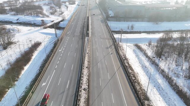 Snow-covered highway with clear lanes shows vehicles traveling in both directions, surrounded by a snowy landscape and roadside infrastructure, captured from an elevated perspective. Aerial view