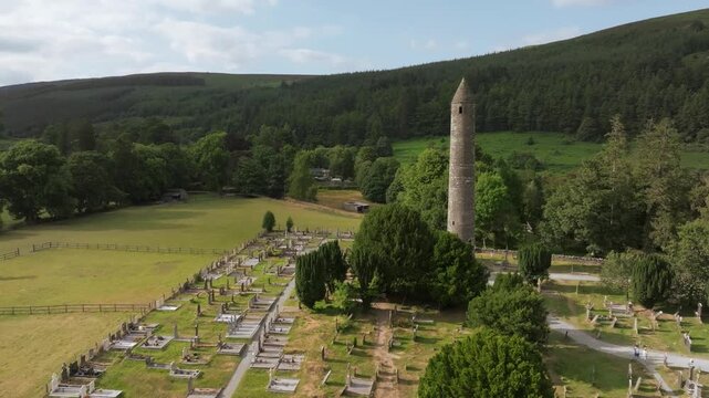 An aerial view shows a tall stone round tower with a conical roof above cemetery paths and rows of gravestones, surrounded by trees, open grassland and wooded hills. Glendalough Co.Wicklow