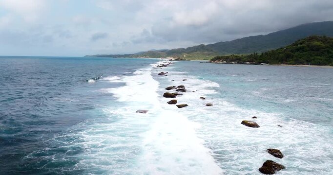 Drone view flying over ocean waves crashing on a long rocky reef with green coastal hills in the background in Tayrona National Park, Colombia