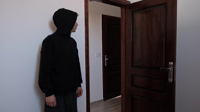 Anxious man in black hoodie hesitating at an open apartment door, struggling to leave home, experiencing fear, uncertainty and emotional distress in a quiet domestic interior