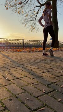 Low-angle vertical video of a female runner stretching and jogging in an autumn park at sunrise. Fitness, healthy lifestyle, morning routines, and outdoor exercise.