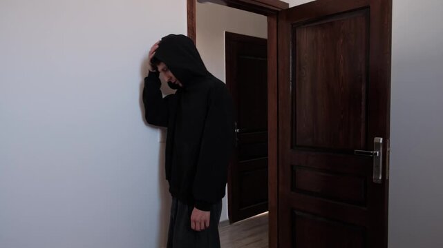 Anxious man in black hoodie hesitating at an open doorway, struggling to leave home, retreating to the wall in fear, avoidance and emotional distress