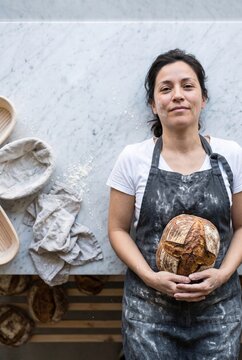 Portrait of a smiling female baker wearing a flour-dusted apron holding a freshly baked artisan sourdough bread loaf in a professional bakery