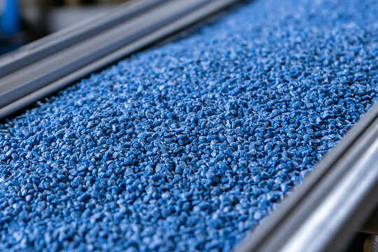 Blue plastic resin pellets move along an industrial conveyor belt. High-tech manufacturing process and polymer materials supply chain concept.
