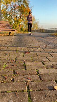 ground-level vertical video showing a woman jogging away from the camera down a paved brick path in a park during autumn.