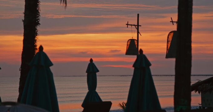 Silhouettes of closed parasols and lanterns on tropical beach at sunset. Calm sea view from luxury resort coast during golden hour. Summer holiday travel concept.