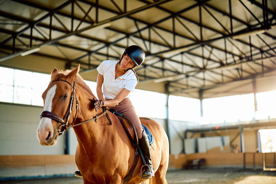 Helmeted woman riding a chestnut horse in an indoor arena, leaning forward and handling the reins during equestrian training