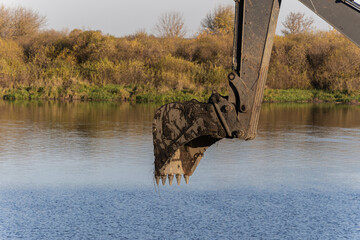 Excavator bucket hangs over calm river water at autumn industrial construction site copy space © Sveta