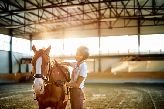 Woman rider standing beside a chestnut horse, checking its saddle in a sunlit indoor riding arena at golden hour
