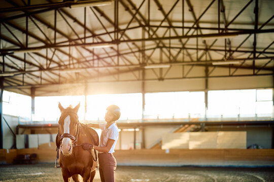 Equestrian standing beside a chestnut horse and holding its reins in a sunlit indoor riding arena, with warm backlighting