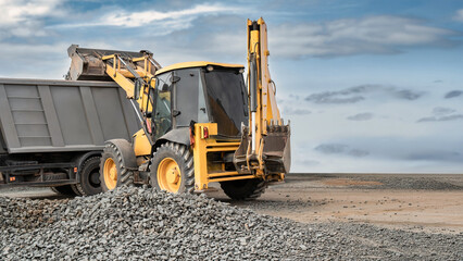 A yellow backhoe loader lifts gravel into a dump truck at a construction site, with piles of stone across the foreground © Anoo