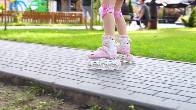 Close up of child legs in pink inline skates and knee pads riding along a alley in a summer park in slow motion. Active kids outdoor leisure concept. Rollerblading.