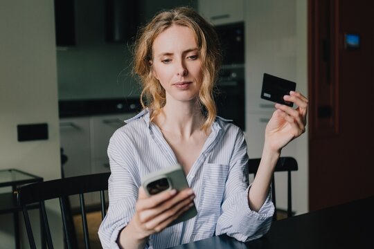 Woman holding a credit card and checking her smartphone at home, illustrating online payment fraud risks, financial scams and the danger of sharing banking information with cybercriminals