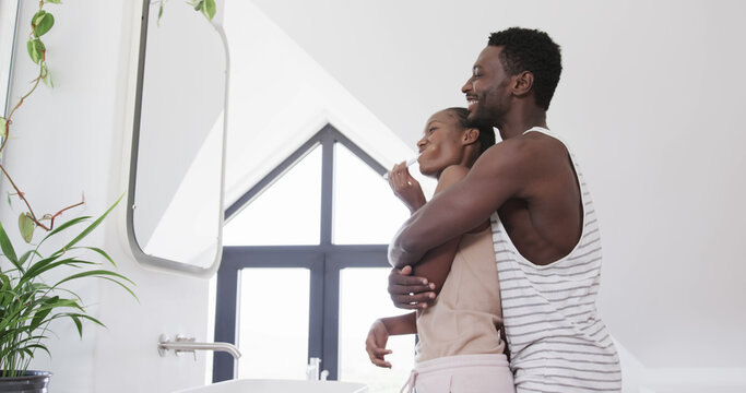 Happy african american couple embracing and brushing teeth in sunny bathroom