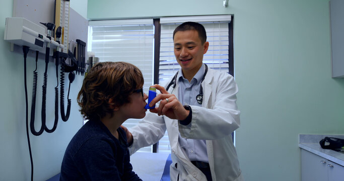 Young asian male doctor helping a caucasian boy patient in using asthma inhaler in clinic