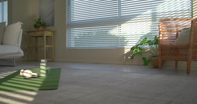 Green yoga mat is basking in sunlight near window blinds while holding water bottle and headphones