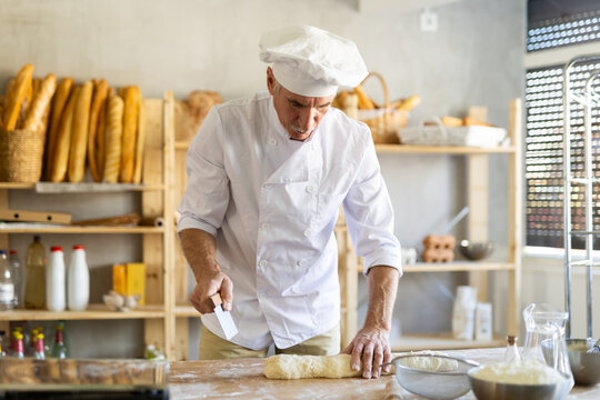 Elderly experienced male baker using knife to cut raw dough to make croissants