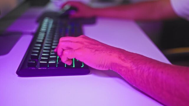 Young man typing on rgb keyboard with hand on mouse at desk in studio under purple led light; focus.