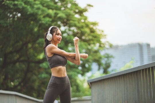 Young Asian athletic woman wearing wireless headphones stretching her arms outdoors in urban park, warming up before fitness workout.