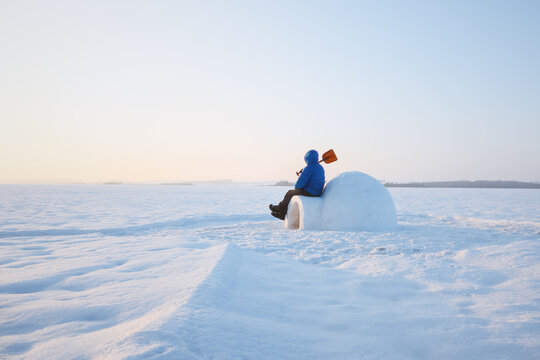 Man in blue winter clothing sit beside a snow igloo on a vast frozen landscape. Blue sky and untouched snow create a peaceful Arctic camping scene