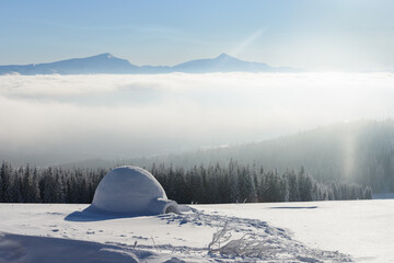 A snow igloo stands on a frozen landscape above a sea of morning fog with distant mountain peaks on the horizon. Soft sunlight and pale blue tones create a serene winter scene © Ivan Kmit