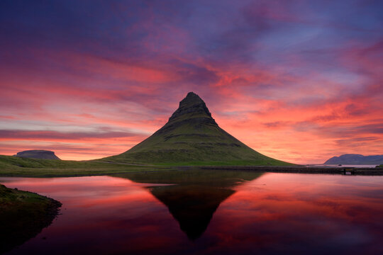 A dramatic volcanic Kirkjufell mountain reflects in a still lake beneath a vivid pink and purple sunset sky. Dark hills and colorful clouds create a striking Icelandic landscape. Iceland