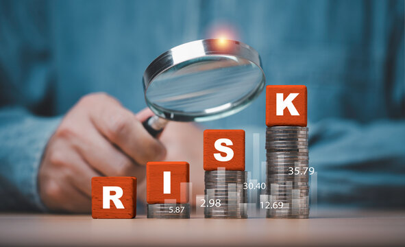 Businessman use magnifying glass inspect to RISK blocks on rising coin stacks and financial charts representing investment risk assessment in volatility market and risk management concept.