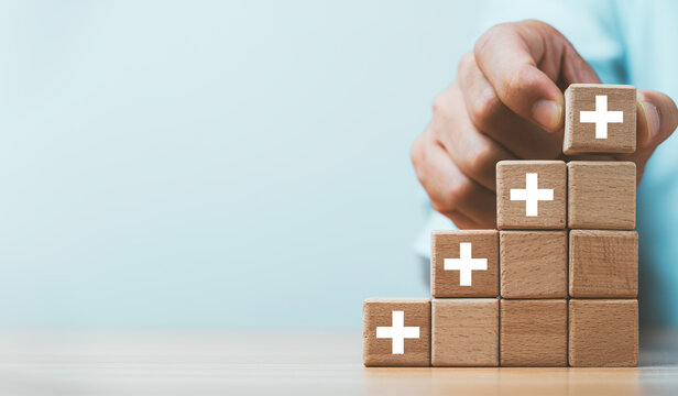 Hand placing a plus sign wooden block on rising steps symbolizing added value for business growth, positive improvement, increasing benefits and continuous development concept.