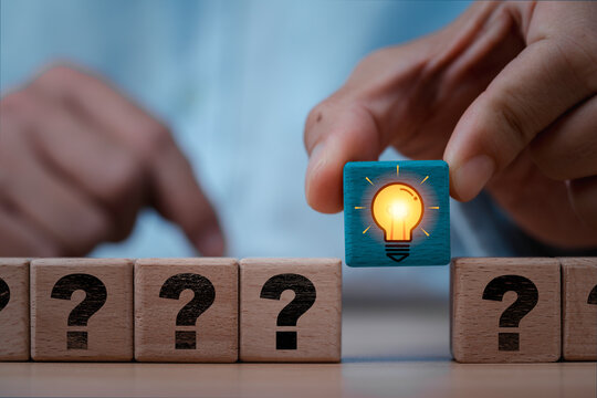 Hand placing a glowing light bulb wooden block among question marks symbolizing creative thinking to problem solving, innovation from bright ideas and inspiration solutions concept.