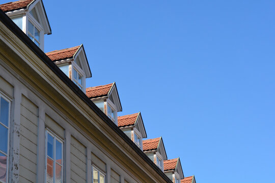 Low angle view of a historic building roof featuring a repeating row of classic white dormer windows with orange clay tiles under a clear blue sky