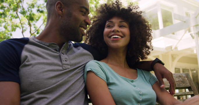 Happy diverse couple sitting on bench talking and embracing in sunny garden
