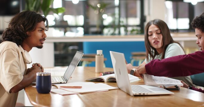 Working three peers, rightmost wearing maroon top, pointing at paper by two laptops in workspace