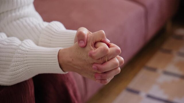 Close-up of a nervous woman fidgeting, rubbing, and wringing her hands together while sitting on a couch indoors.