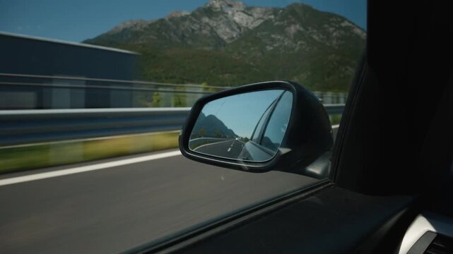 Driving on highway past side mirror reflecting asphalt road and alpine mountains near A23 Udine.