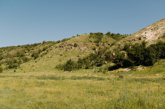 wide grassy meadow leading to sunlit rocky hillside under clear blue sky, gentle summer light, distant shrubs