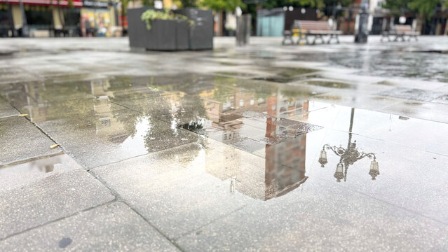 Rainwater puddles reflecting city buildings and ornate street lamps across wet stone pavement in an urban square after rainfall