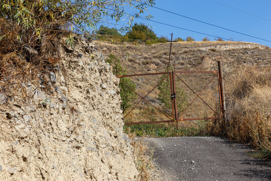 Closed rusty metal gate locked with padlock on gravel road near dirt hill in rural area