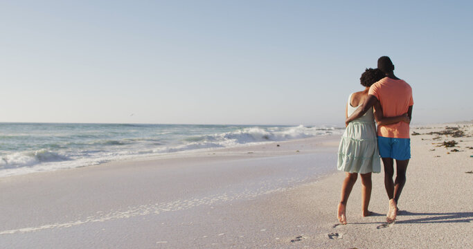 African american couple embracing together and walking on sunny beach