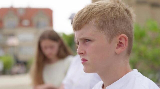 A sad boy looks at the camera in the background his family mother and sister are sitting on a bench