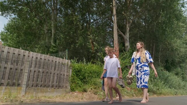 Woman and two children are walking barefoot along a tree lined road family walk the boy is biting his fingers