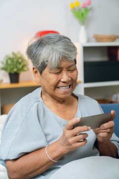 Elderly Asian woman relaxing on a sofa at home, enjoying games on her smartphone, modern living among older adults.