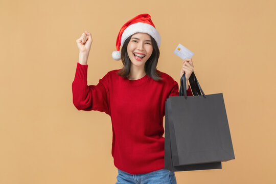 Cheerful Asian woman celebrating Christmas wearing a santa hat and holding a credit card with shopping bag while smiling, black friday online shopping and modern lifestyle concept.